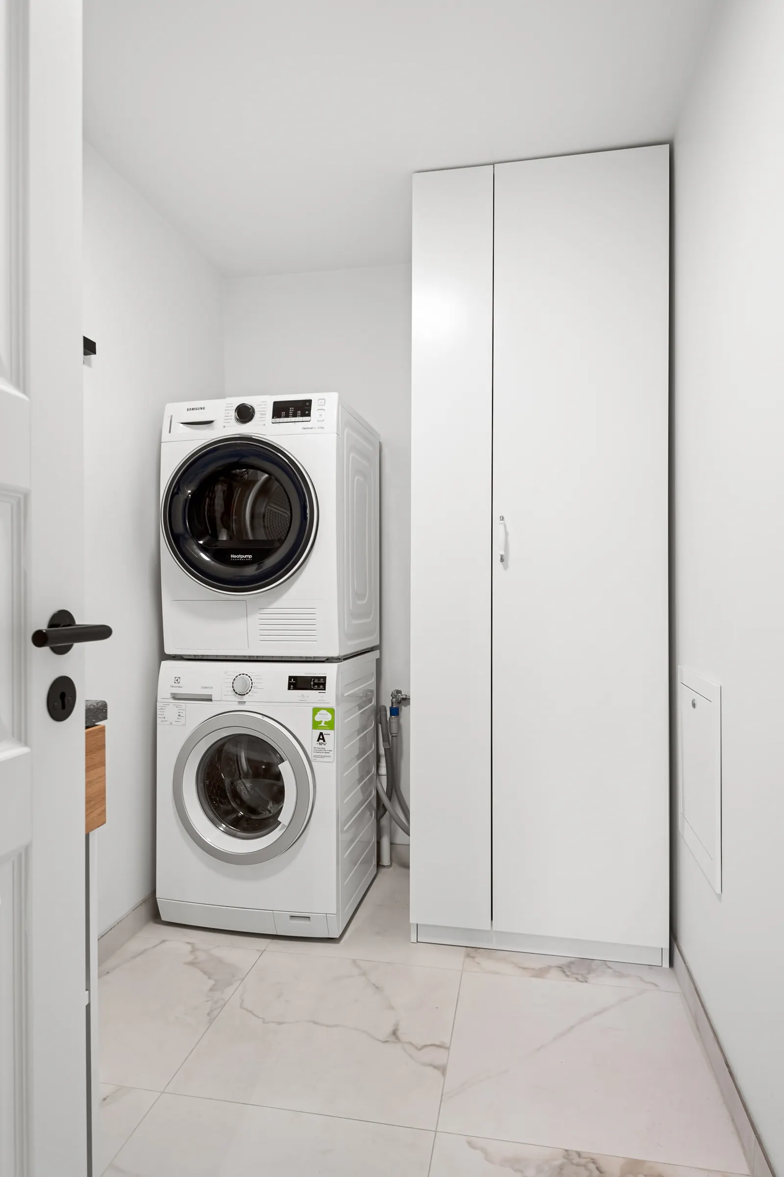 Stacked washer and dryer beside a tall storage unit in a bright narrow laundry room